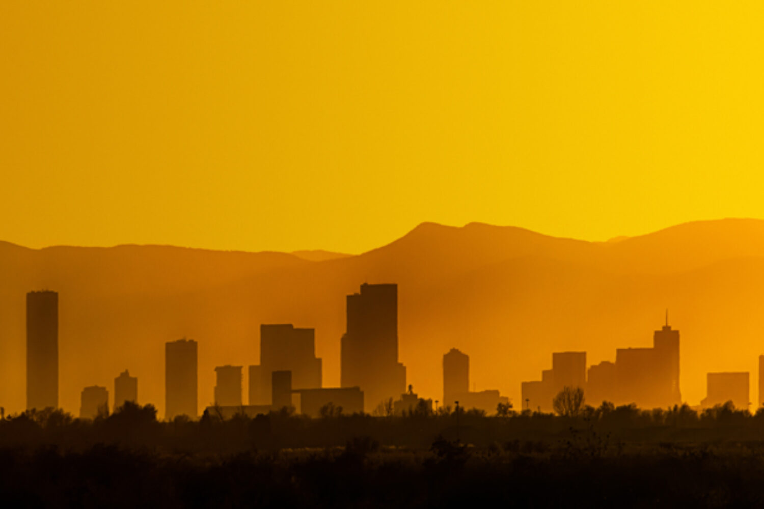 A bright and hazy orange sunset silhouettes the Denver, Colorado skyline against the Foothills of the Rocky Mountains.