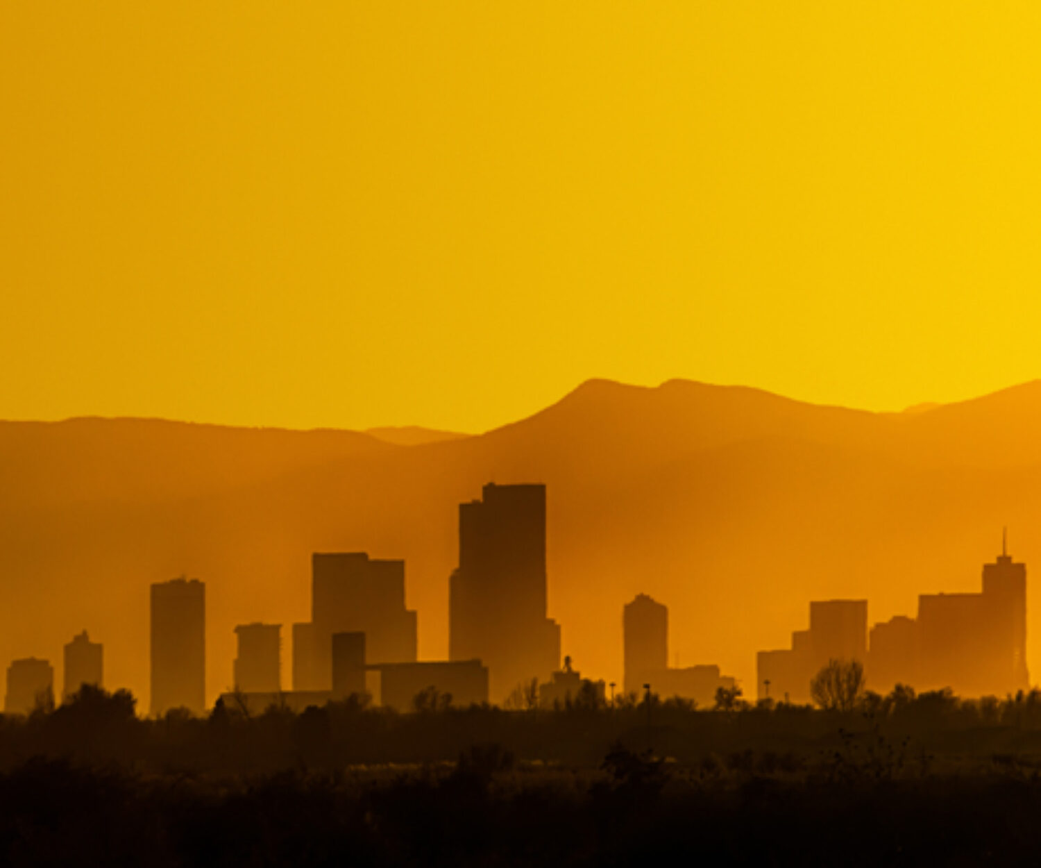 A bright and hazy orange sunset silhouettes the Denver, Colorado skyline against the Foothills of the Rocky Mountains.
