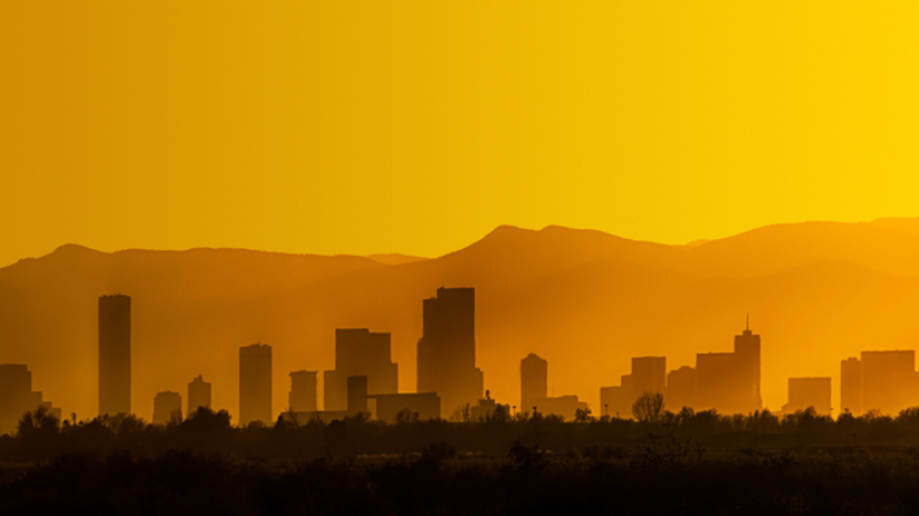 A bright and hazy orange sunset silhouettes the Denver, Colorado skyline against the Foothills of the Rocky Mountains.